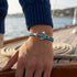 Maritimes Ankerarmband für Herren mit türkisem Seil vor dem Meer getragen