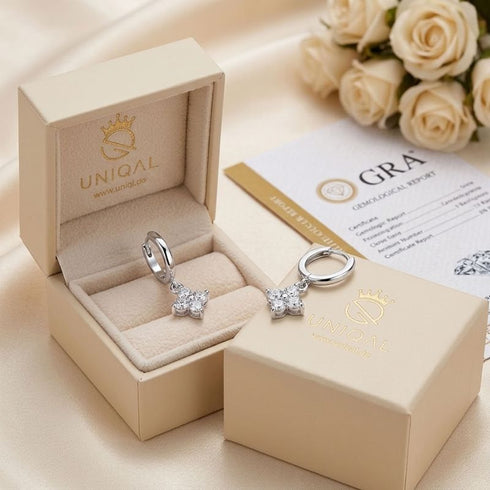 ÉCLAT von UNIQAL als Geschenkarrangement mit cremefarbener Schmuckbox, einem Ohrring im Etui und einem auf dem Deckel, neben weißen Rosen und GRA-Dokument.