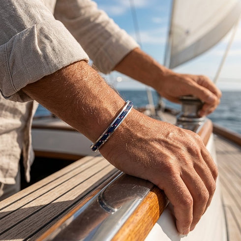 UNIQAL MONACO Edelstahl-Armreif am Handgelenk eines Mannes mit beigem Hemd an Bord eines Segelboots.