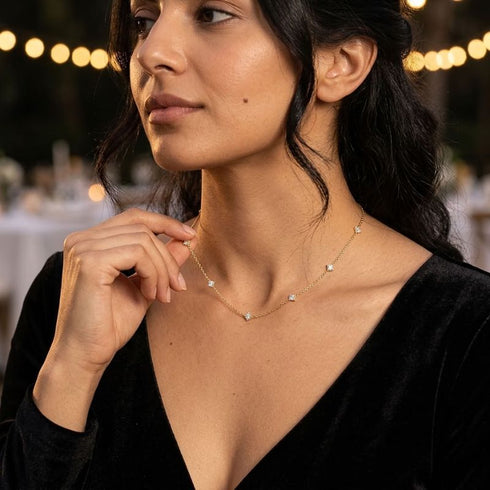 SHINE CELESTA 14K GOLD von UNIQAL ergänzt einen schwarzen V-Ausschnitt mit funkelnder Halszier, aufgenommen vor einer stimmungsvollen Lichterkulisse.