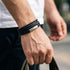Uniqal BLACK OLIVE Armband am Handgelenk – schwarzes mehrreihiges Herrenarmband mit geflochtenen Details und silberfarbener Metallplatte für einen lässigen Outdoor-Look.