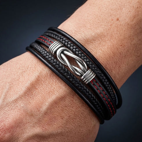 ENDLOS von UNIQAL am Handgelenk: mehrreihiges schwarzes Lederarmband mit glatten und geflochtenen Bändern, roten Nähten und silberfarbenem Infinity-Motiv.