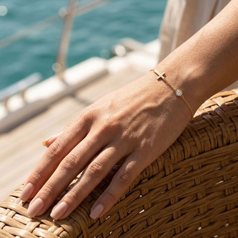 GRACE 14K GOLD Moissanit-Armband von UNIQAL an einer Hand auf einem geflochtenen Korbsessel, im Sonnenlicht vor türkisfarbenem Wasser