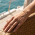 GRACE 14K GOLD Moissanit-Armband von UNIQAL an einer Hand auf einem geflochtenen Korbsessel, im Sonnenlicht vor türkisfarbenem Wasser