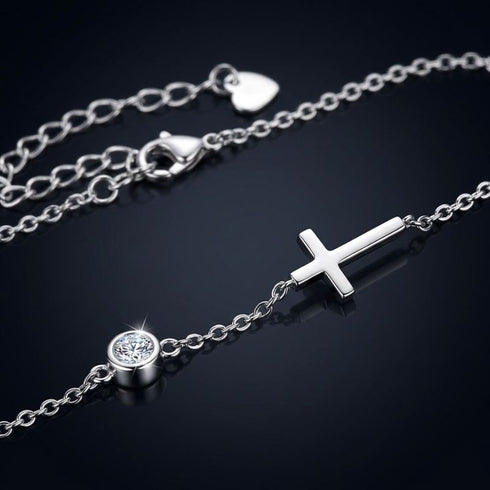 UNIQAL GRACE Moissanit-Armband auf dunkler Oberfläche, mit poliertem Kreuzmotiv, Karabinerverschluss und kleinem Herzplättchen am Kettenende.