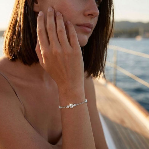 Frau mit GRAZIA Moissanit-Armband von UNIQAL zu einem beigefarbenen Oberteil, die Hand an der Wange im warmen Sonnenlicht am Hafen