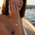 Frau mit GRAZIA Moissanit-Armband von UNIQAL zu einem beigefarbenen Oberteil, die Hand an der Wange im warmen Sonnenlicht am Hafen